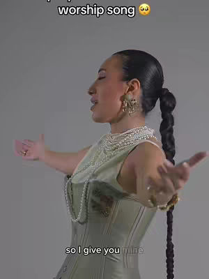 A woman stands with arms outstretched, appearing to praise or worship. She wears a corset-style top, adorned with multiple layers of pearl necklaces and large gold earrings. Her dark hair is styled with a long braid, and she's set against a neutral background. Text overlays are at the top and bottom of the frame.
