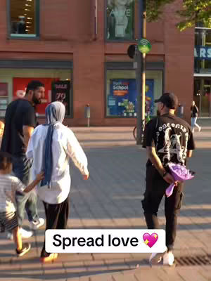 The cover image shows a street scene with several people. The foreground features a person wearing a blue hijab walking with a child holding their hand. Behind them, a man with a beard is visible, and to the right, another man is seen from the back; he is dressed in black with a cap, carrying a purple bouquet. Buildings in the background.