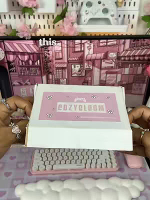 A person holds a white box with a pink label reading "COZYBLOOM" over a pink and white setup including a keyboard, mouse, and a decorative cloud. The background features a light pink aesthetic.
