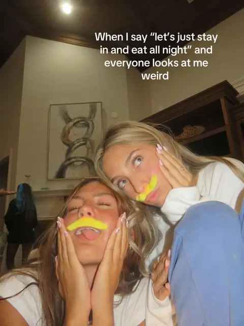 A selfie-style image featuring two young women indoors. They each have a bright yellow mustache drawn on their upper lip, and their hands are positioned near them. Text overlayed on the image says: 'When I say 'lets just stay in and eat all night' and everyone looks at me weird.' The lighting in the room is soft and warm.