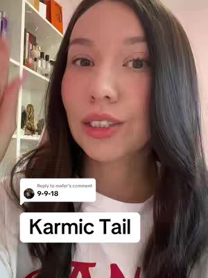 A woman with long dark hair is centered in the frame, looking directly at the camera. She wears a white top. A comment reply with a date is overlaid on her image, and a banner with text reads 'Karmic Tail'. The background includes a partially visible white shelf with various items.