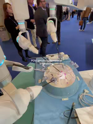 The image shows a surgical simulation setup with robotic arms in a clinical/exhibition hall setting. The overhead view captures a blue, circular medical bay or testing area. Robotic arms, resembling sleek white tools, are positioned above a model or patient simulator. The scene suggests a demonstration or training environment focused on technological advancements in medicine.