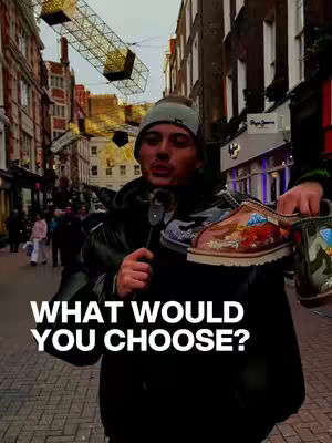 A man stands on a city street, holding shoes and a microphone. The background shows buildings and a street scene.