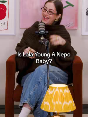 A woman is seated in a chair, angled towards the viewer, with a microphone in front of her. The background includes abstract artwork. Text over the image asks, "Is Lola Young A Nepo Baby?"