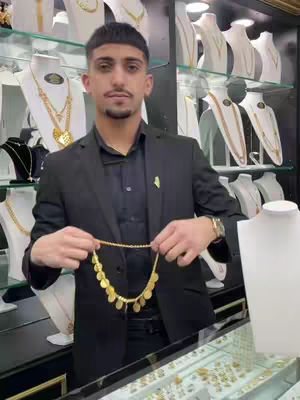 A man in a black suit holds a gold necklace in a jewelry store. The background displays various gold necklaces on display stands. The overall lighting is bright, highlighting the gold jewelry.