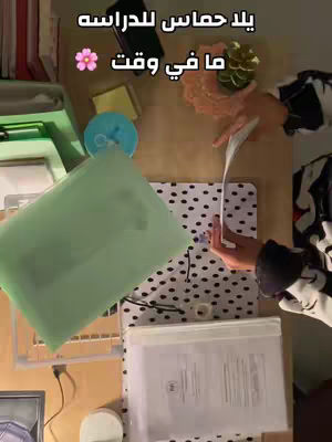 An overhead shot emphasizes a study setup. A light-colored desk has various items like books, green folders, a blue container, and a polka-dotted pad. A hand writing or highlighting is center right. Text overlays in Arabic appear at the top. The composition uses natural light, suggesting a casual, active study environment.