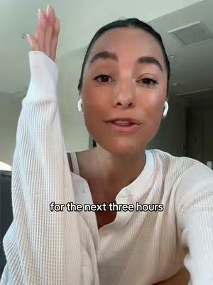 A woman with light skin and dark hair is shown in a close-up shot. She is wearing a white shirt and has white earbuds in her ears. Her right arm is raised with her palm facing the viewer, suggesting she may be gesturing. She has a friendly expression. The background is blurred, with a ceiling fan or light fixture partially visible.