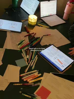 A top-down shot of a table filled with study materials: multiple laptops and tablets displaying documents, scattered papers, pens, and a lit candle.