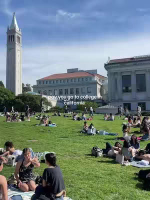 An outdoor scene on a college campus, featuring a large grassy area filled with students. Buildings, including a tall tower and other structures, form the backdrop under a blue sky.