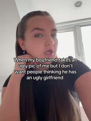 A young woman with long brown hair is facing the camera, against the background of a bright room. She's wearing a black top and appears to be talking. Text overlayed on the image reads, "When my boyfriend takes an ugly pic of me but I don't want people thinking he has an ugly girlfriend."