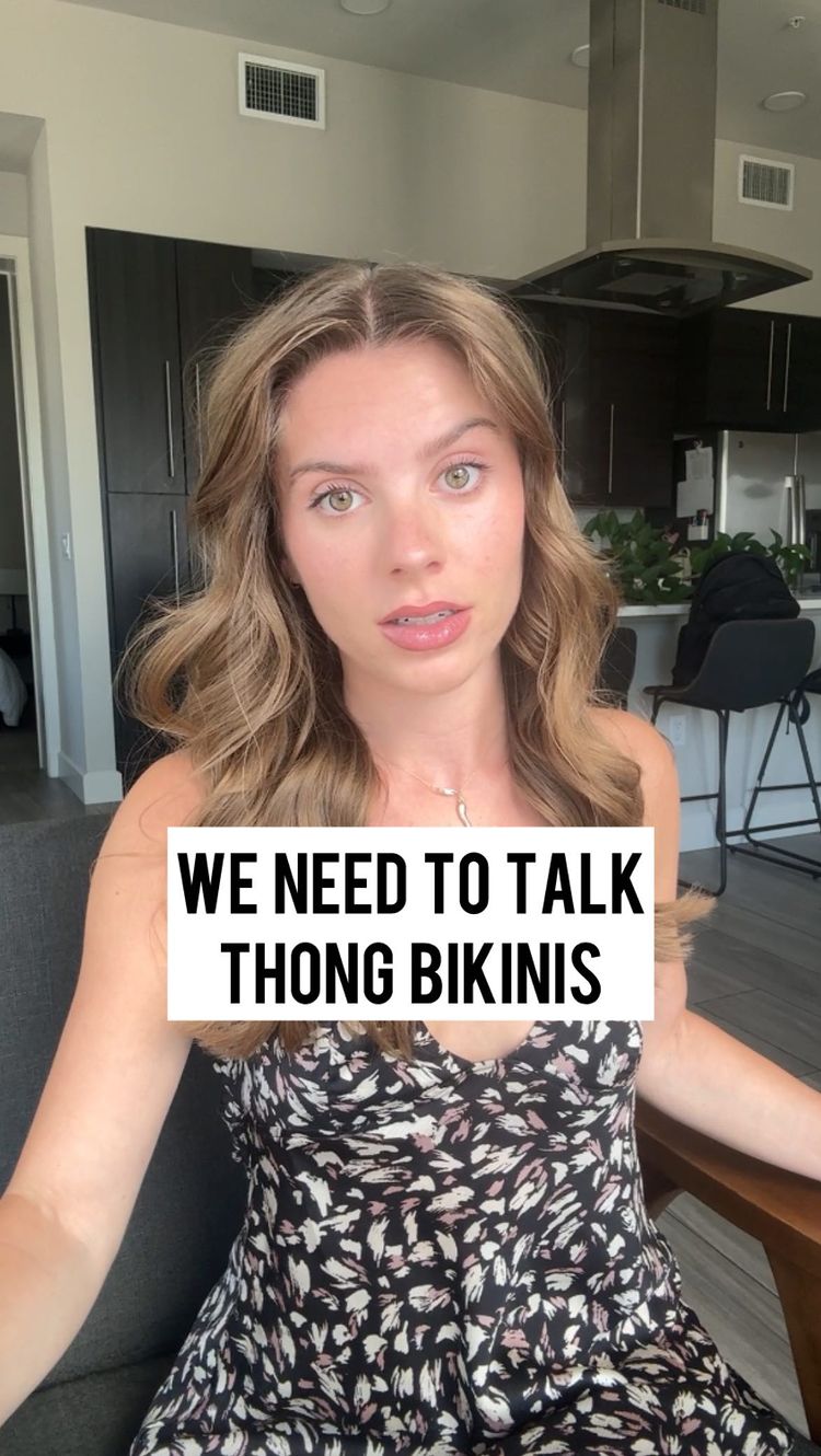 A woman with long, wavy brown hair is centered in the frame, looking directly at the camera. Above her is a text box that reads "WE NEED TO TALK THONG BIKINIS." She is wearing a black and white floral print dress. The background shows parts of a kitchen.