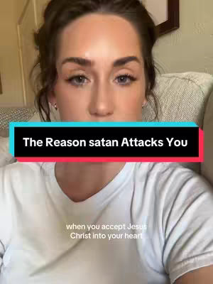 A woman with brown hair and fair skin is centered in the frame. She wears a white t-shirt and looks directly at the viewer with a neutral expression. The background is out of focus, and a text overlay in a black and red box reads, "The Reason satan Attacks You," positioned above her chest. Small, light text appears below, stating "when you accept Jesus Christ into your heart."