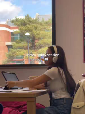 A young woman with long dark hair wearing a white t-shirt and headphones sits at a wooden desk in a library. A laptop is open in front of her. She is facing the computer with her hand near the trackpad and there are books on the desk. Behind her, a large window shows a view of trees, a building, and the sky. The overall composition is well-lit, focusing on a studious scene.
