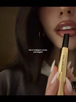A close-up shot shows a woman holding a golden tube of lip gloss near her mouth. Her lips are glossy and prominent, and she has long, manicured nails. Text overlaid says, 'she is intelligent, classy and elegant'.