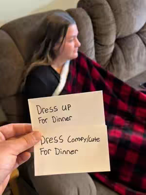 A person is holding a note in front of a seated woman on a brown couch. The note has handwritten text: 'Dress Up For Dinner Dress Comfy/cute For Dinner.' The seated woman is slightly out of focus, wearing a black shirt and a red and black plaid blanket. The background includes a brown couch.