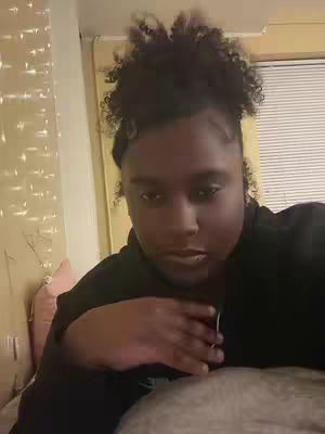 A woman with dark curly hair is sitting, facing the camera with her hand touching her chest; she's wearing a black top and is positioned in a modestly lit room. The background includes a window with closed blinds, and a hint of a decorative element or fairy lights to the left.
