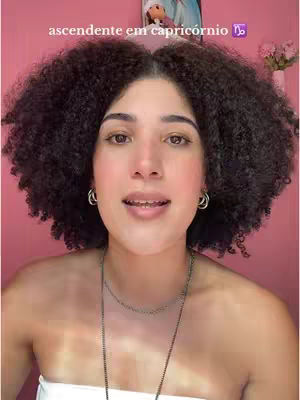 A woman with curly dark hair is centered in the frame, looking at the camera with a neutral expression. She wears a white tube top and a necklace. The background is a solid pink color. Above her head is a painting in a frame. Text overlay at the top says 'ascendente em capricórnio'.