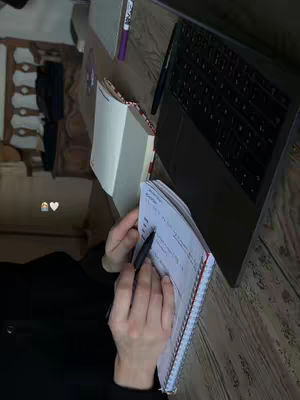 A person's hands writing in a notebook, with a laptop and other stationery items visible on a wooden desk. The image has a slight warm tone, enhanced by the lighting, and the overall composition suggests a study or work setting.