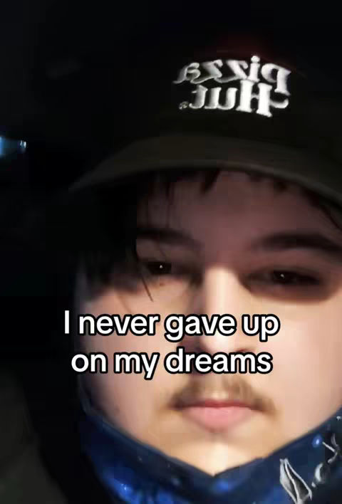 A close-up selfie shows a young man wearing a dark cap, with text overlayed. The background is very dark, suggesting a low-light setting. The man has a mustache; the text reads 'I never gave up on my dreams'.