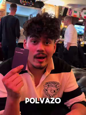 A young man with curly hair holds up a card from a board game, with the game's title overlaid at the bottom. He appears surprised with his mouth open. In the background, there are people, seemingly in a pub.