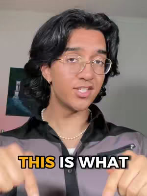 A young man with dark hair and glasses is centered in the frame, pointing downwards. He's wearing a gray and black shirt and a pearl necklace. A painting can be seen in the background. Overlaying the bottom of the image are the words "THIS IS WHAT".