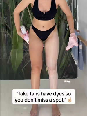 A person in a black bikini applies self-tanner to one leg, showing the contrast between the tanned and untanned skin. A white text box overlays with the text "fake tans have dyes so you don't miss a spot". Tropical looking foilage forms the backdrop and a pink glove is set to hang on a towel rack.