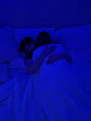 A dimly lit image of a couple embracing in bed. The scene is suffused with a deep blue hue. Two figures are visible, one with dark hair, nestled close to the other person, who has their back to the camera. They are under white bedding, with pillows adding to the cozy setup, and the angle suggests an intimate and relaxed atmosphere.