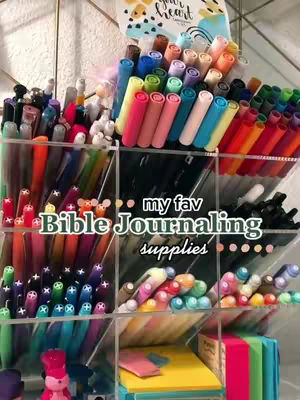 A visually organized display of pens, markers, and other stationery items. The items are neatly arranged in transparent acrylic containers to create a colorful and aesthetically pleasing image. The composition is well-lit, highlighting the variety and vibrancy of the supplies, implying a focus on Bible journaling.
