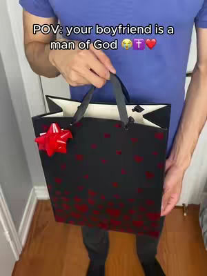 A person, likely male, stands holding a large black gift bag decorated with red heart-shaped patterns, secured with black handles, and a red bow. The person is wearing a blue collared shirt, and the scene takes place indoors against a neutral backdrop.