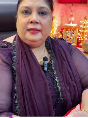 A woman with dark hair is the focal point of the image. She is facing the camera, with the upper half of her body visible, seated. She has a fair complexion and is wearing dark eye makeup and red lipstick and a dark purple outfit. A microphone is visible on her chest. The background is softly lit with decorative elements. The overall composition suggests a focus on the subject within a studio setting.