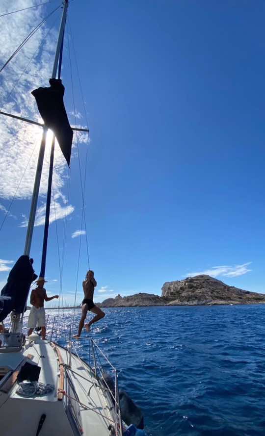 Several people are on a sailboat, with a man jumping into the sea. The sky is blue with some scattered clouds, and there's a small island in the distance. The scene is bright and sunny.