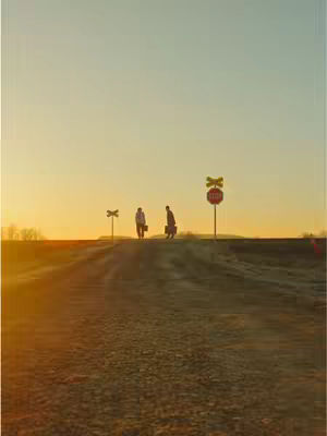 A wide, outdoor shot taken at sunset or sunrise. Two figures, silhouetted against the bright sky, walk away from the camera along a slightly sloped path. They carry briefcases or similar bags. Railroad crossing signs and a stop sign punctuate the horizon, and the overall composition is balanced with warm, golden hues.