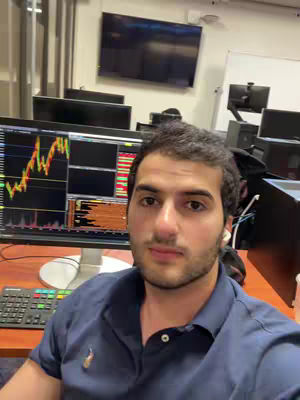 A man is in a well-lit office setting. The man, who appears to be the focus of the frame, is sitting at a desk and looking directly at the camera. Behind him are multiple computer monitors displaying financial data, with charts and numbers. The office setup includes a keyboard, desk, and other office equipment, with a large TV screen on the wall. The overall composition suggests content related to finance or investing.