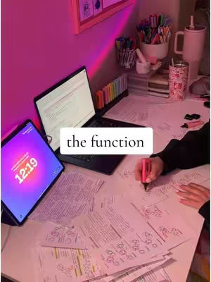An overhead shot of a desk setup for studying, with a laptop and tablet displaying content, surrounded by papers and stationery, bathed in a pink light.