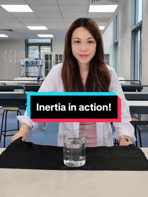 A woman in a lab coat stands behind a table. On the table is a beaker of water and a black cloth. The text overlay 'Inertia in action!' is in the center of the image.