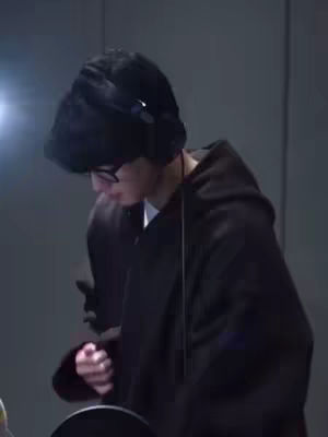 A man wearing glasses, headphones and a black hoodie is in a recording studio, with his head bowed down.