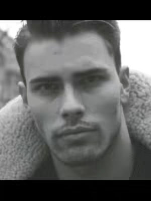 A grayscale close-up portrait of a man. He gazes directly at the viewer with a neutral expression and close mouth. The background is blurred, and the man has short, dark hair and a slight mustache. He is also wearing what appears to be a fleecy, light-colored jacket.