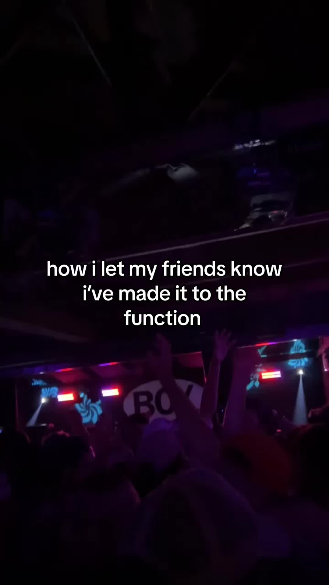 The image is a low-angle shot from inside a crowded, dimly lit venue, likely a concert or club. Colored lights, predominantly red and blue hues, illuminate the scene. The foreground shows partially visible figures with raised hands. In the mid-ground, text is overlaid in white, set against the darker backdrop. Towards the back, a blurry outline of a stage and more figures suggest a performance.