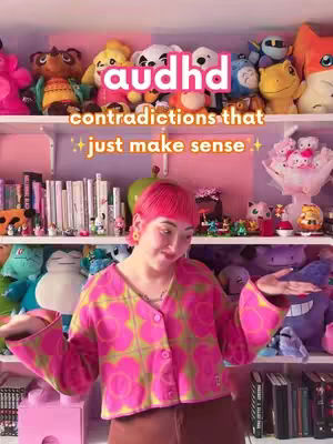 A person with bright pink hair and a pink floral patterned cardigan stands in front of a shelf filled with plush toys and books. The background features additional shelves with various decorations.