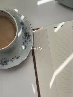 An overhead shot of a white surface with a cup of coffee on a floral plate, accompanied by an open book. Sunlight casts shadows, enhancing the scene's aesthetic.