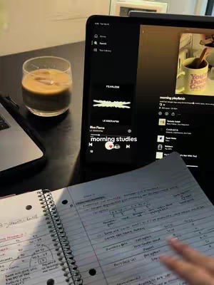 A well-lit image of a study desk setup. A clear glass of iced coffee sits on the left, next to a laptop. In the center, a tablet displays a music app with a playlist titled 'morning studies'. A notebook with notes and handwritten text is in the foreground, with a hand placed on the page. A mug with text on it is visible at the top right corner.