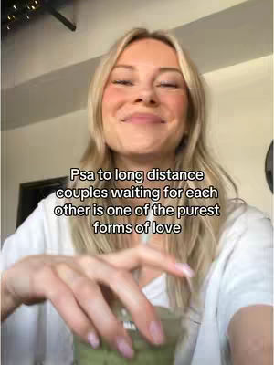 A woman with long blonde hair is smiling slightly, holding a green drink in a clear cup. Overlaid text reads a PSA about long-distance couples. The background appears to be an interior setting with a bit of a dark corner.