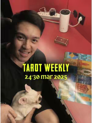 A man with dark hair smiles while seated, holding a small dog. The background is a red desk with a power strip and a white accessory. Text overlay in bright yellow indicates the video's content.