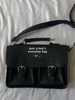 The image displays a black, rectangular bag with a flap, buckles, and a shoulder strap, set against a white, textured background, likely fabric. The bag is the primary focus and positioned centrally. Text overlaid on the bag adds context.