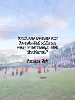 The image shows a sunny outdoor scene with a green grassy field. In the background, there are several people, some wearing various colored shirts and playing what looks like a game. Dominating the center are the words in white of a bible verse set over the background, and the sky is light blue with a slight gradient effect.