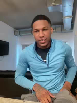 A medium shot shows a Black man facing forward and slightly angled toward the right. He has short, dark hair and a neatly groomed appearance. He's wearing a light blue, long-sleeved shirt over a black t-shirt, and a tattoo is visible on his left arm. The background shows a modern setting with visible air ducts and a flat-screen TV.