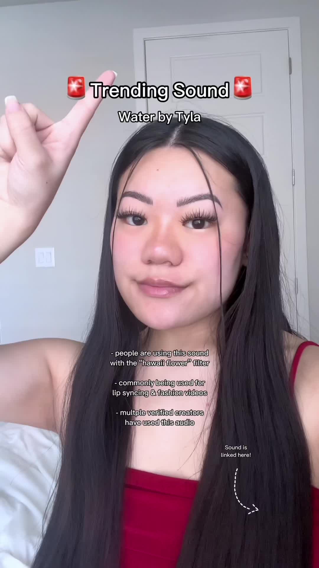 A woman with long, dark hair is pointing upwards with her right hand, almost as if she's touching something above her head. Her face is visible, demonstrating makeup and a soft expression. The background is a plain wall with a door partly visible.