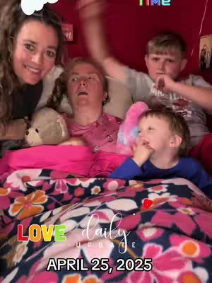 A selfie-style video cover featuring a group of people in a bedroom setting. It includes four individuals: a woman on the left smiling, a woman in the middle lying down, and two young boys present in the frame. They are laying on a bed, covered with a vibrant floral blanket, and the room's red background suggests an intimate setting. Text overlays with dates and messages like 'LOVE daily UPDATE' are visible. Overall it conveys a sense of familial closeness and a special moment.