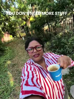 A woman with reddish-brown hair and oversized eyeglasses is holding a blue coffee mug. She is smiling and facing the camera. The background is a blurred mix of green foliage, giving a natural, outdoor setting.