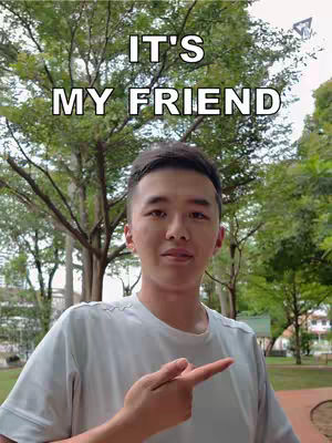 A young man with short dark hair stands in front of a blurry green background of trees and foliage. He is wearing a light blue t-shirt and pointing to the right with his right index finger. A text overlay, written in a white, bold font, reads "IT'S MY FRIEND" above the man.